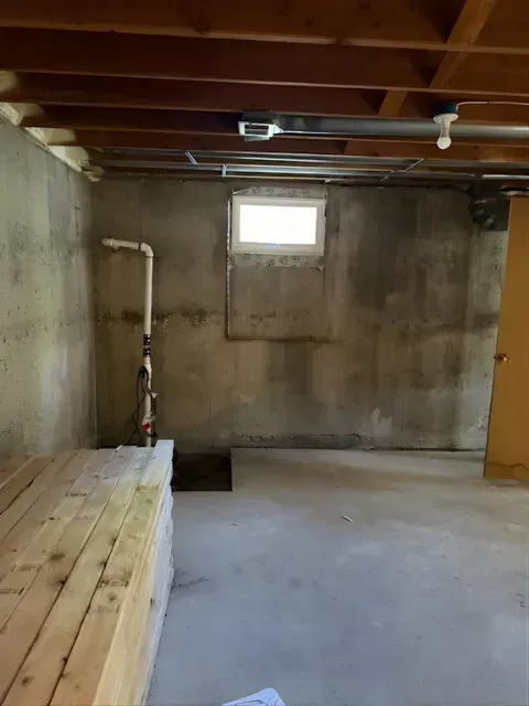 Unfinished basement with concrete floor, exposed ceiling beams, small window, and wooden platform along the left wall