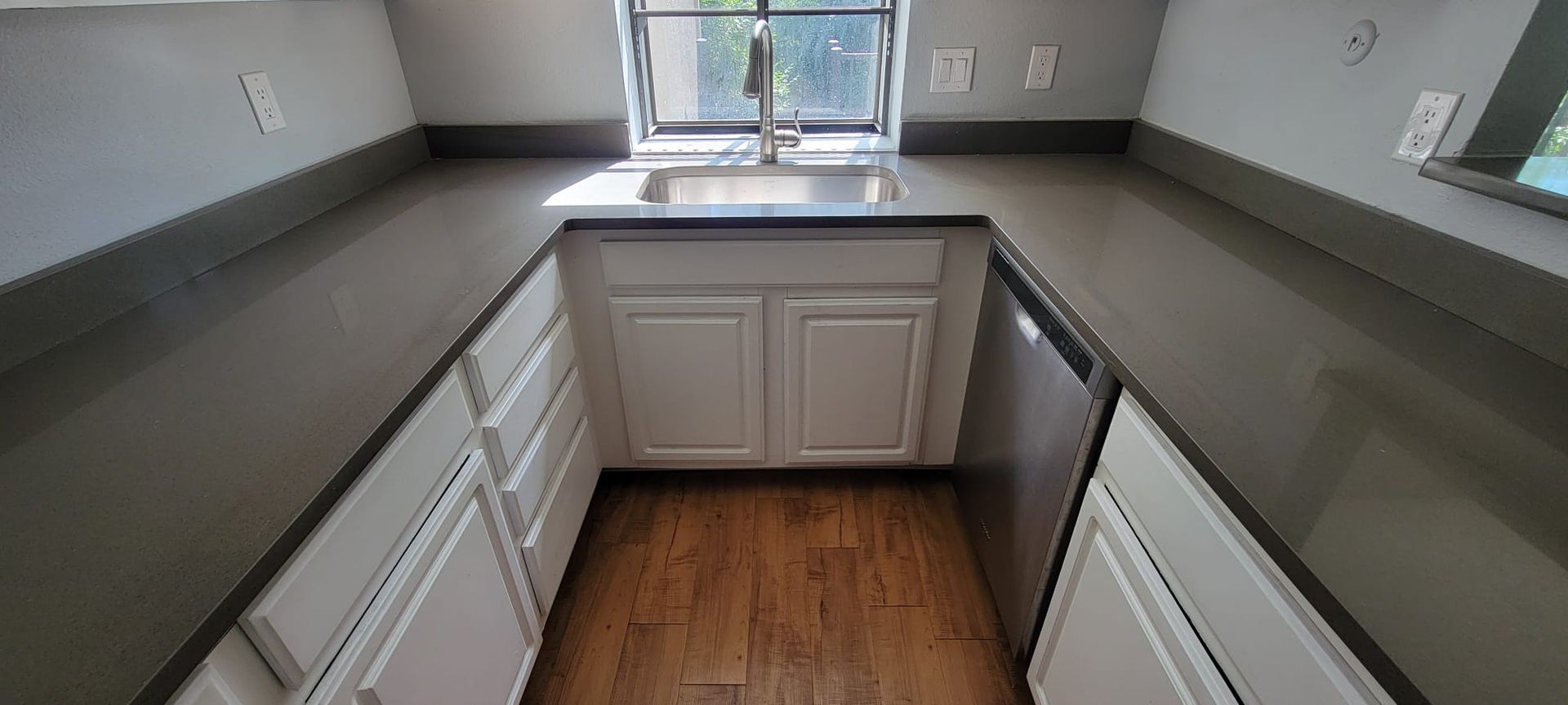 Empty kitchen with gray counters, white cabinets, wood floor, and a sink under a window.