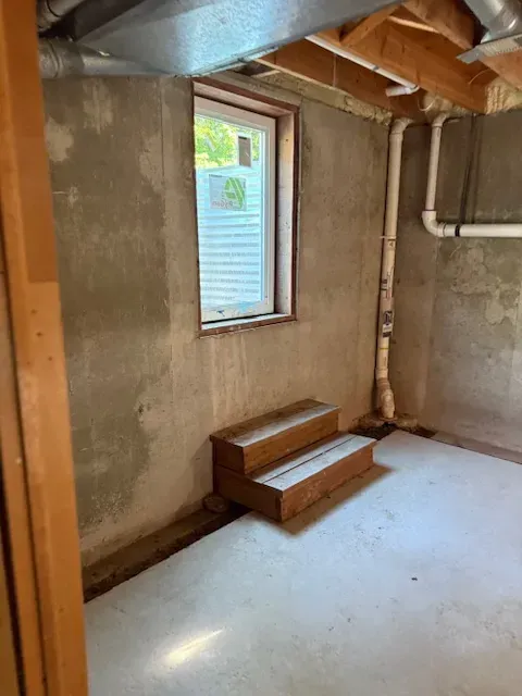 Bare unfinished room with concrete walls, exposed beams, and a small window above two wooden steps.