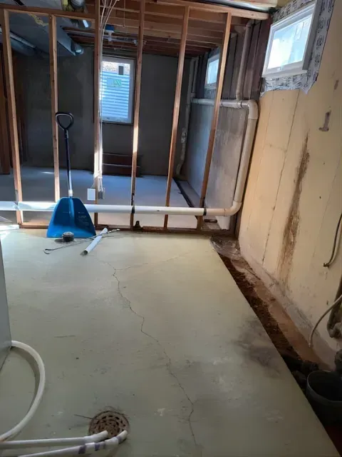 Partially renovated room with exposed framing, white pipes, and a blue dustpan on a bare concrete floor