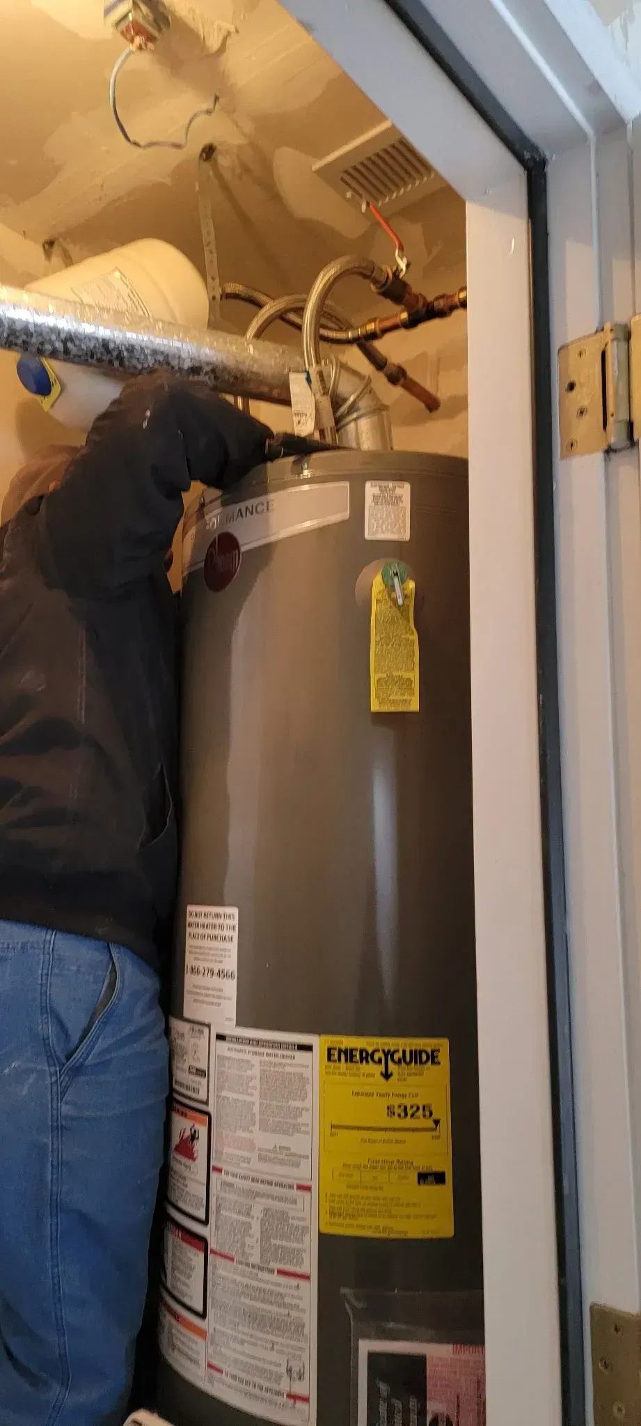 Technician in a closet working on a gray water heater with exposed wiring and pipes.