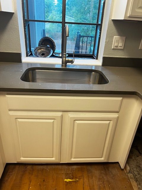 Kitchen sink beneath a window, with white cabinets and dark countertop on a hardwood floor.