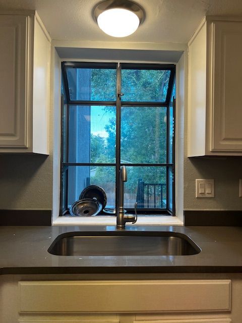 Kitchen sink beneath a window with a forest view, framed by light cabinets and a ceiling light above.
