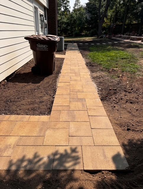 Yellow paver walkway along a house beside mulch beds and trash bins, leading toward a yard