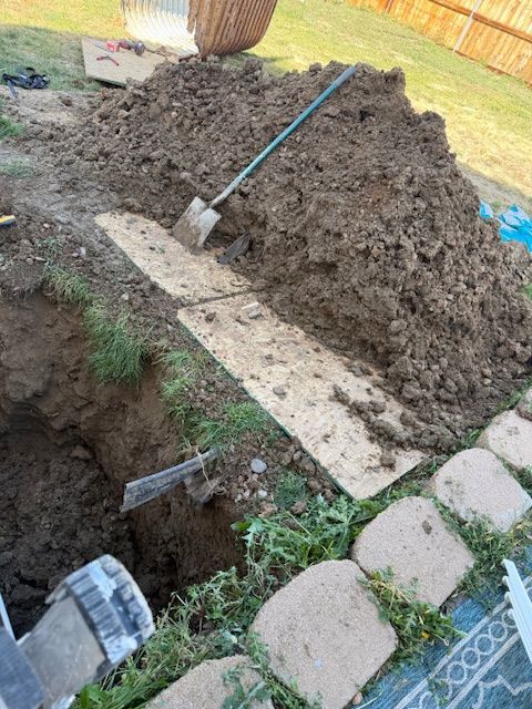 A shovel in a dirt pile beside an open trench next to a brick walkway and gray utility boxes