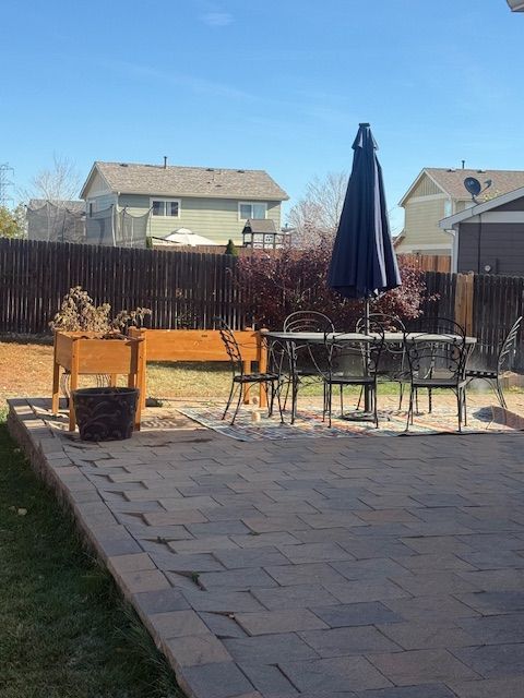 Backyard patio with black metal table, umbrella, planter boxes, and fenced yard under a blue sky