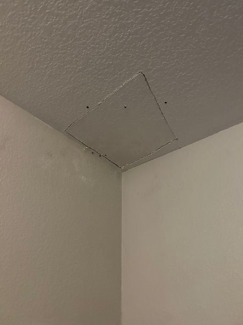 Corner of a ceiling with a small square access panel and visible screws, painted light gray.