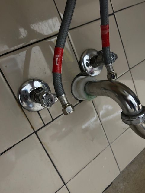Chrome faucet plumbing against beige tile wall with two flexible hoses and round wall valves