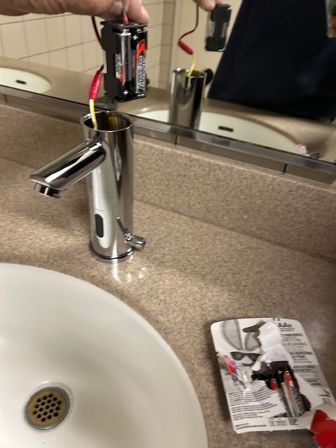 Hand holding a 9V battery above a bathroom sink faucet, with packaging on the counter
