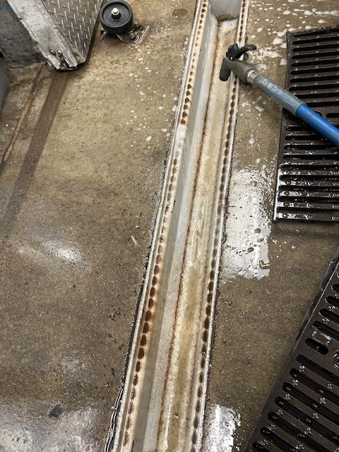 Close-up of a wet concrete floor beside a drain grate and blue hose in an industrial area