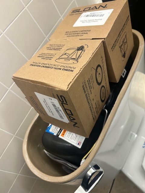 Cardboard box resting on top of a black trash can in a bathroom.
