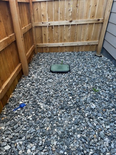 Gravel-floored fenced yard with a green utility cover and a small blue object on the left.