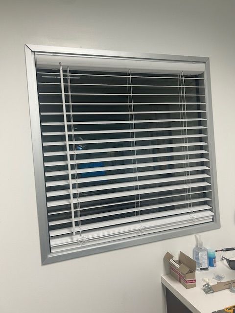Window with white blinds and a desk corner with office supplies beneath it.