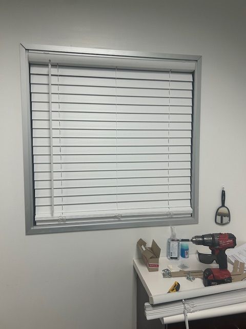 A small window with white blinds above a white desk holding tools and a drill in a work area.