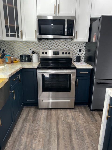 Compact modern kitchen with stainless steel appliances, dark cabinets, white backsplash, and gray wood flooring