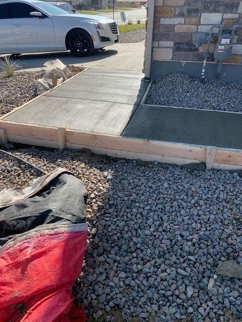 Front walkway under construction beside a stone house, with gravel, concrete slabs, and a parked car nearby