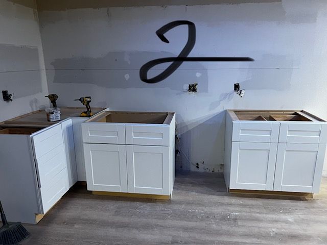 Kitchen under renovation with white cabinets and gray flooring, sink openings visible on unfinished counters