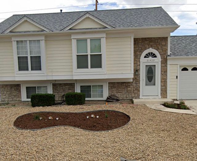 Suburban townhouse with beige siding, gray roof, front door, and gravel yard with a small mulched bed