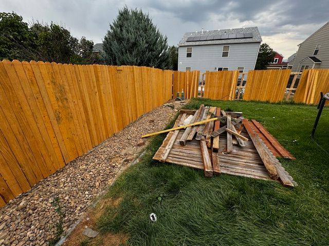 Backyard with wooden fence, gravel path, and piled lumber on a grassy area under cloudy sky