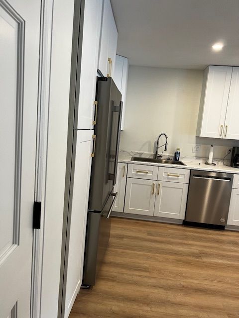 Modern kitchen with white cabinets, stainless steel appliances, and wood flooring, viewed from a hallway