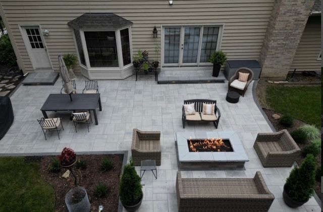 Patio with outdoor sofa, chairs, fire pit, and dining table on a stone terrace beside a house.