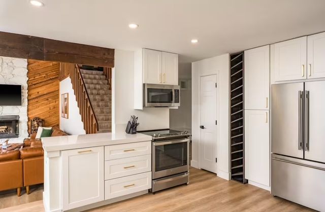 Modern white kitchen with stainless steel appliances, wood flooring, and a view into a living room.