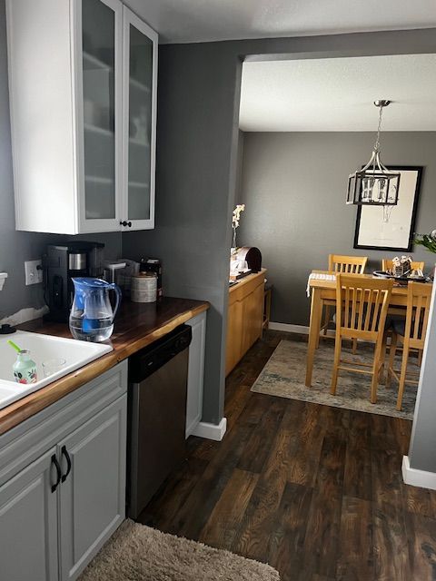 Kitchen looking into dining room with gray cabinets, dark floor, and wooden table under a hanging light.