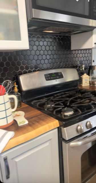 Kitchen stove with stainless steel oven, black hexagon tile backsplash, and wooden countertop with mug and utensils.