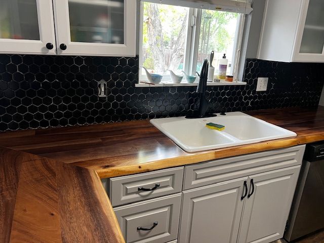 Kitchen sink beneath window with black hex tile backsplash, wood countertop, and white cabinets