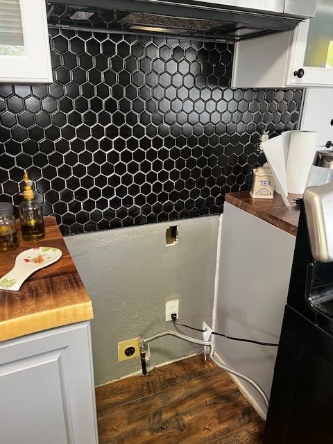 Small kitchen corner with black hex tile backsplash, wooden floor, and countertop items near a stove.