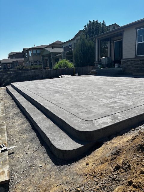 Concrete patio slab under construction beside suburban homes, with raised edges and dirt around it