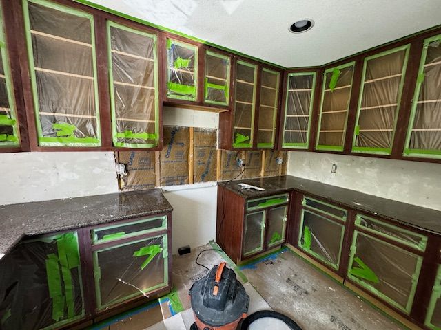 Kitchen cabinets being painted dark brown with green masking tape; floor protected by a vacuum and drop cloth.