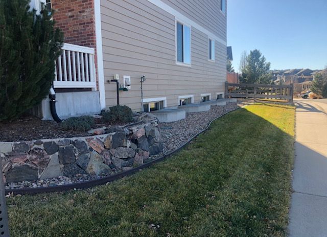 Side yard of a beige house with stone retaining wall, green lawn, and concrete sidewalk beside a sunny road