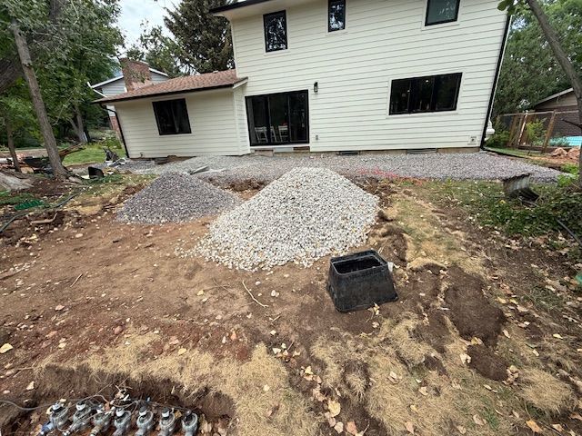 Backyard under construction beside a white house, with dirt piles, gravel mounds, and a black tub.