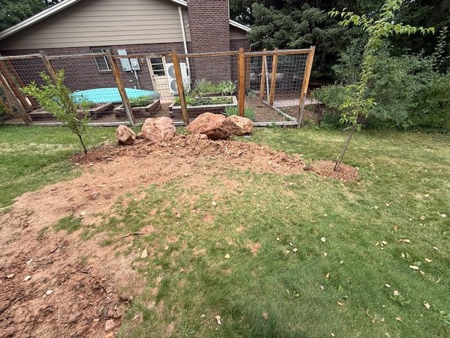 Backyard with freshly dug dirt mound, rocks, and a wooden fence beside a house and trampoline