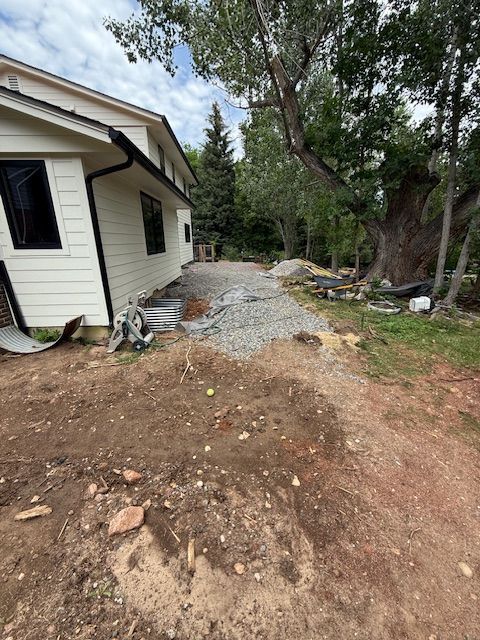 Side yard beside a house with gravel, dirt, trees, and landscaping debris.
