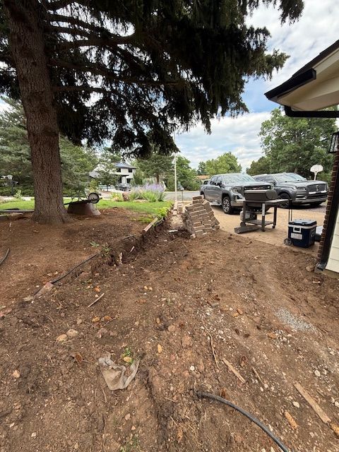 Dirt trench beside a house with parked cars, large trees, and a cloudy sky in a residential yard