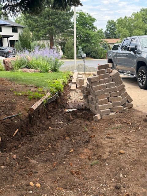 Driveway construction site with stacked pavers, excavated dirt, and a parked SUV beside a lawn.