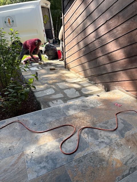 Person working on a stone walkway beside a brown house, with an orange extension cord in the foreground