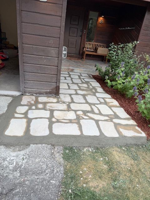 Stone walkway leading to a cabin entrance, bordered by mulch and shrubs, with a bench inside the porch.