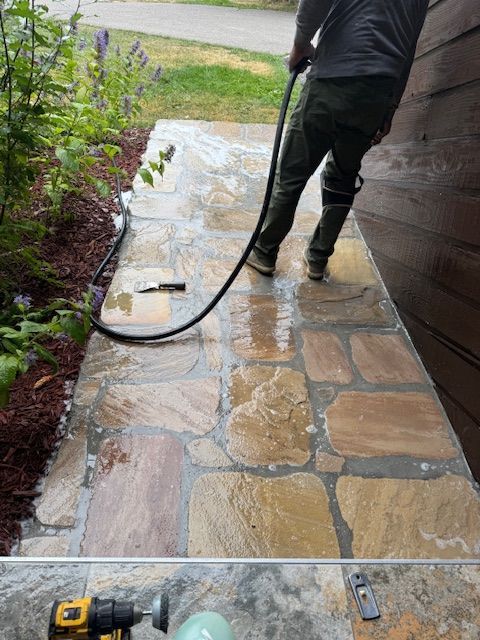 Person pressure-washing a stone walkway beside a house, with wet pavement and a hose visible.