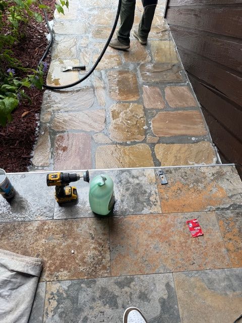 Stone patio under repair with tools, a green bottle, and a worker’s boots visible near a hose.