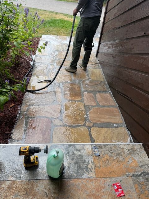 Person pressure-washing a stone walkway beside a house, with cleaning supplies in the foreground.