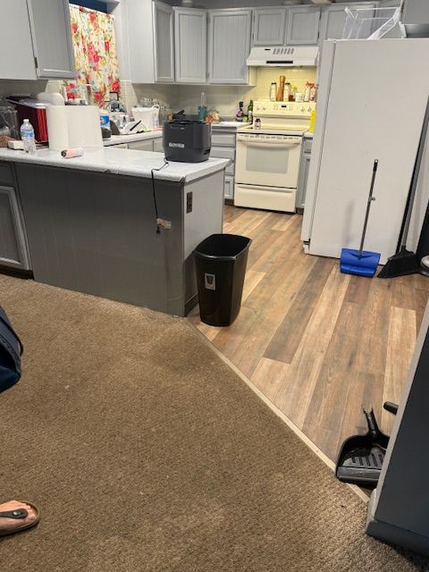Messy kitchen and living area with black trash bin, white cabinets, and brown carpet