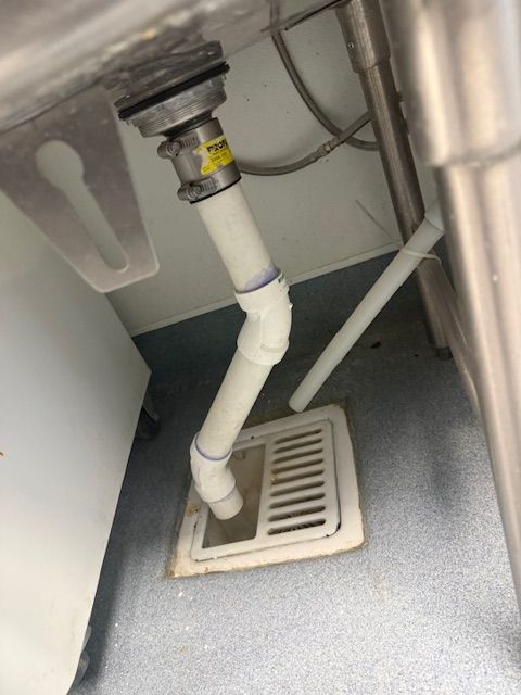 Under-sink plumbing with white drain pipe leading to a floor drain grate on a gray floor