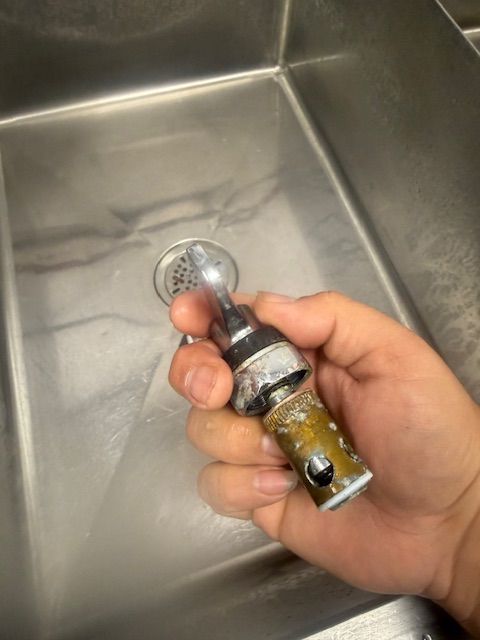 Hand holding a small metal pipe over a stainless steel sink with a drain.