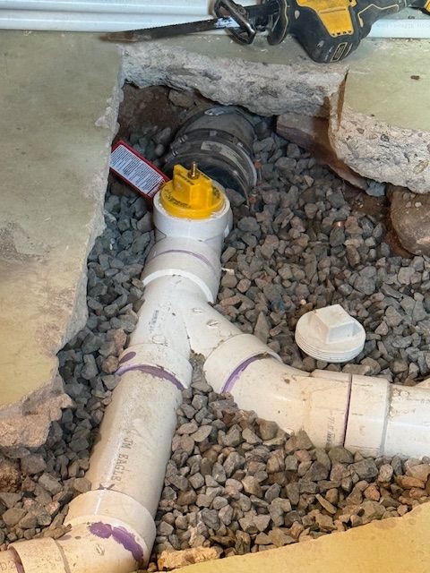 Open plumbing trench with PVC pipes, gravel fill, and a yellow capped fitting under a broken concrete slab