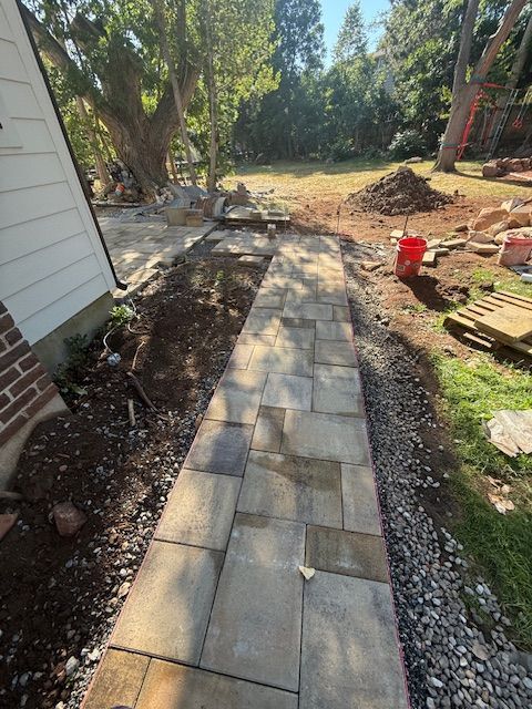 Stone paver walkway beside a house, bordered by gravel and mulch in a sunny backyard.
