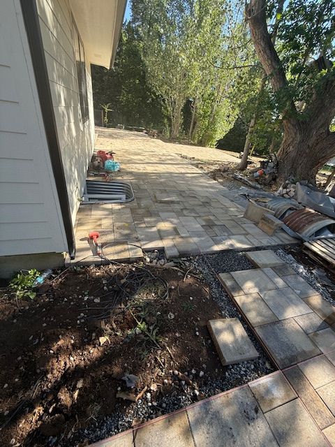 Stone patio beside a house, with a dirt planter, trees, and stacked pavers in a sunny yard.