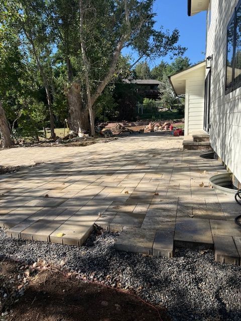 Paver patio beside a house, with trees and gravel edging in a sunny backyard.